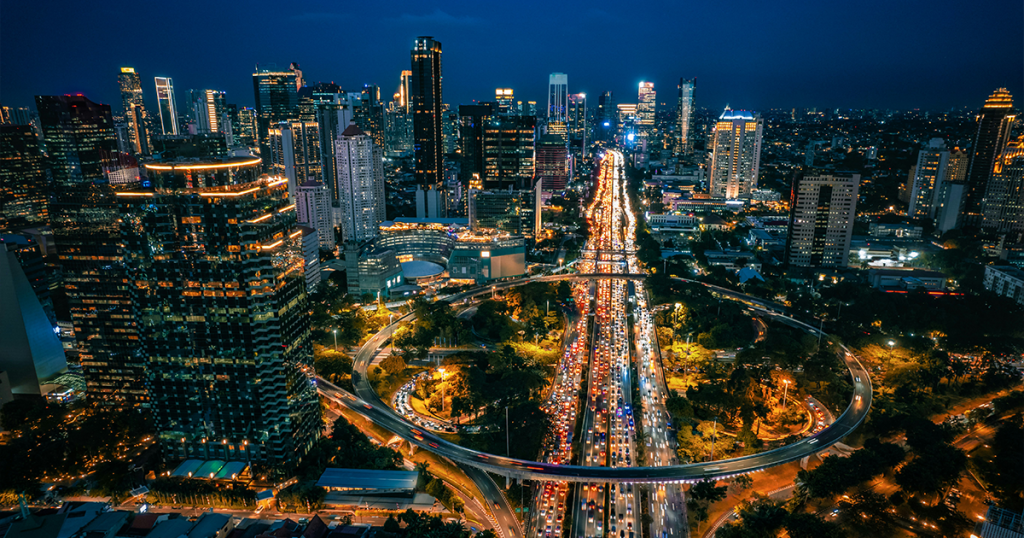 Night aerial of Jakarta showing the Simpang Susun Semanggi interchange with heavy traffic, surrounded by illuminated high-rise buildings.